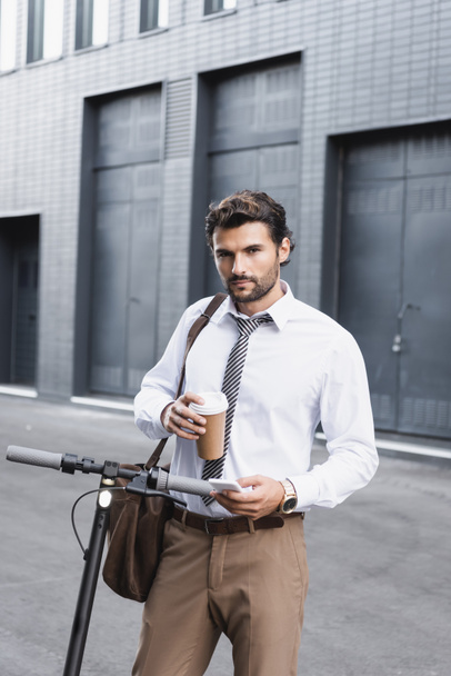 бородатий бізнесмен у костюмі, який тримає каву, щоб піти і використовувати смартфон біля електронного скутера
  - Фото, зображення