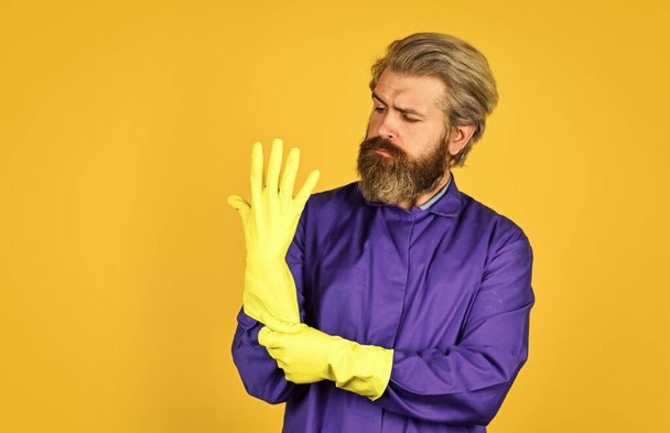 Ochrana. Muž nosí gumové rukavice. Vousatý čistič v modré uniformě žluté pozadí. Jarní úklid. Hipsterský úklid. Dezinfekční koncept. Odstranění viru a mikrobů. Virová dezinfekce - Fotografie, Obrázek