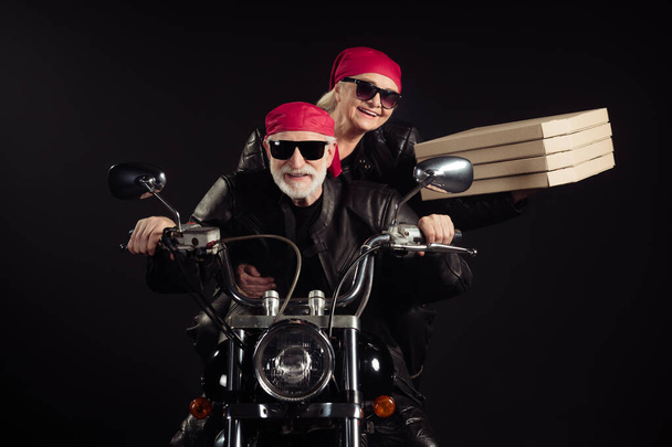 Photo of two cool old bikers white hair man lady couple drive vintage chopper travel festival bring takeaway pizza wear rocker leather jacket outfit bandana isolated black color background - Photo, Image
