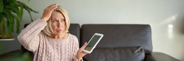 грустная и депрессивная зрелая женщина сидит на диване и с помощью цифрового планшета - Фото, изображение