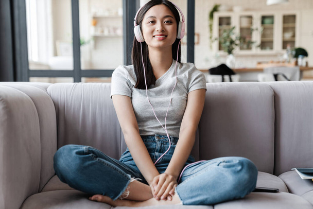 Image of happy asian woman smiling and listening music with headphones while sitting on sofa at home - Fotoğraf, Görsel