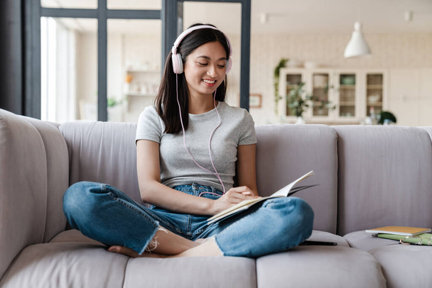 Image of happy asian woman using headphones and writing down notes while sitting on sofa at home - Foto, Imagem