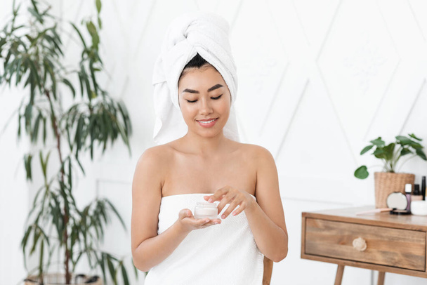 Sorrindo asiático jovem senhora envolto em toalha usando creme - Foto, Imagem