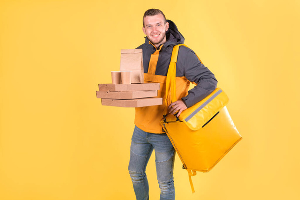 Cute caucasian food delivery courier smiles and looks ahead, holding food order in his hands and yellow refrigerator bag on yellow background hangs on his shoulder. Food delivery to home or office. - Фото, изображение