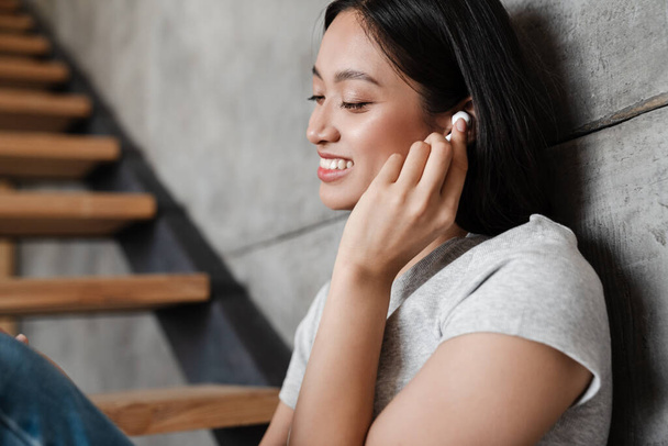 Image of smiling asian student girl using wireless earphones while sitting on stair at home - Foto, imagen