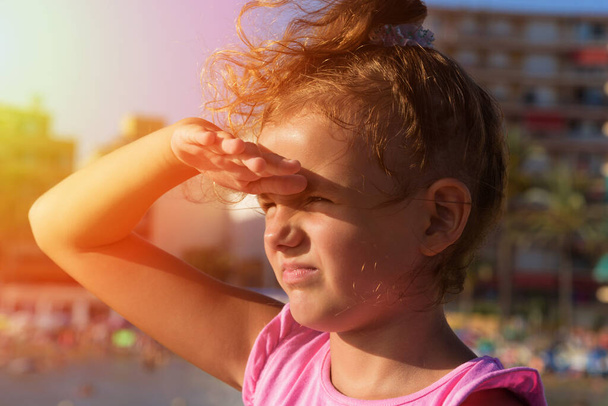Una niña bonita mira lejos de derecha a izquierda, entrecerrando los ojos y jugando al mono bajo el sol en el fondo de la playa de la ciudad. Puesta de sol - Foto, imagen