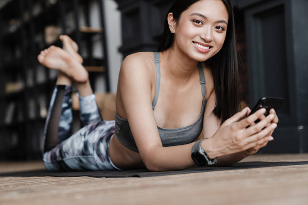 Image of cheerful asian girl in sportswear using mobile phone while lying on yoga mat at home - Fotó, kép