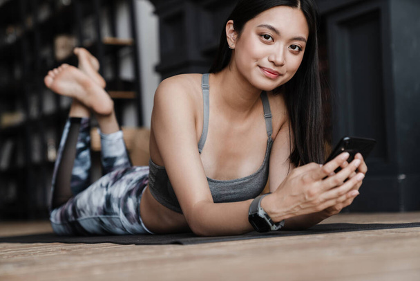 Image of cheerful asian girl in sportswear using mobile phone while lying on yoga mat at home - Fotografie, Obrázek