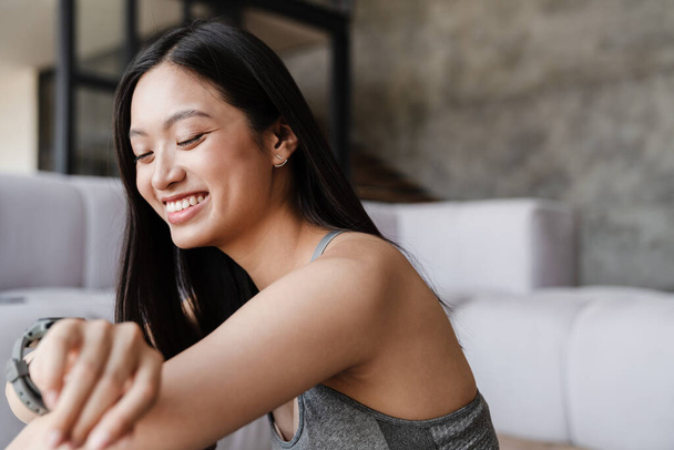Image of happy asian woman smiling and looking at smartwatch while sitting on floor at home - Foto, Imagen