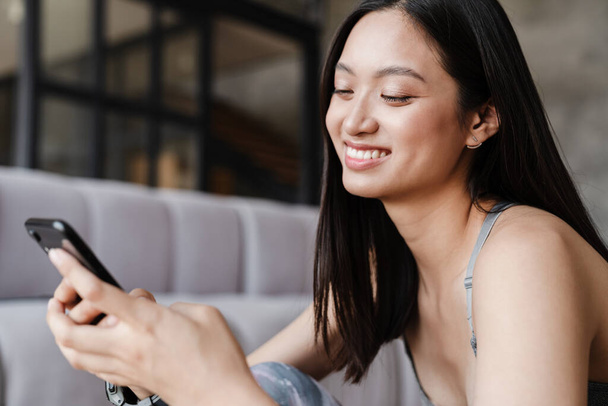 Image of happy asian woman smiling and using mobile phone while sitting on floor at home - Foto, Imagen