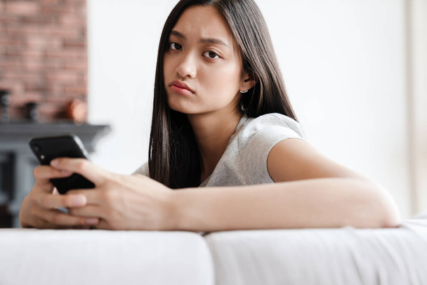 Image of unhappy asian woman using mobile phone while sitting on sofa at home - Zdjęcie, obraz