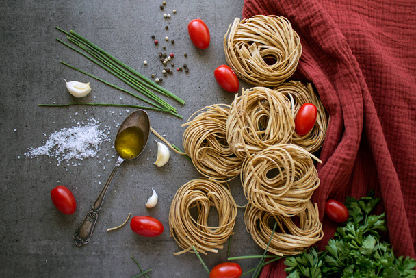Άψητα ζυμαρικά Tagliatelle, σκόρδο, αλάτι, ελαιόλαδο, πιπέρι, ντοματίνια και μαϊντανό σε γκρι πλακάκια τραπέζι κουζίνας. Ιταλικές αποδείξεις φαγητού. Φωτογραφία πάνω όψης των συστατικών ζυμαρικών. - Φωτογραφία, εικόνα