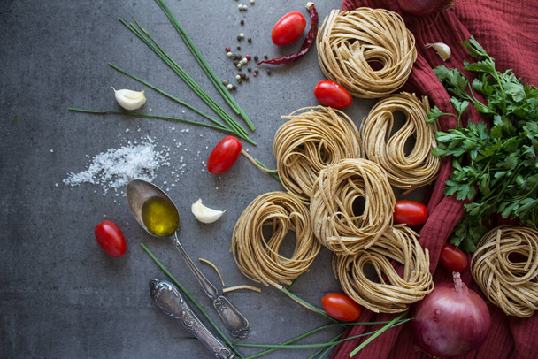 Uncooked Tagliatelle pasta, garlic, salt, olive oil, pepper, cherry tomatoes and parsley on gray tiled kitchen table. Italian food receipts. Top view photo of pasta ingredients. - Photo, Image