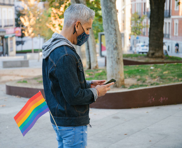 マスクの穏やかな男性の側面ビューとLGBTの虹の旗がマドリードの通りに立っており、コロナウイルスパンデミックの間に携帯電話を閲覧 - 写真・画像