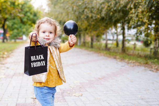 Smiling kid girl holding shopping bag on black friday and black inflatable ball outdoors - Photo, Image