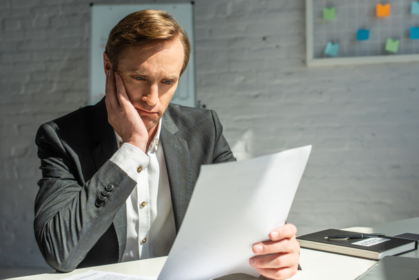 Грустный бизнесмен смотрит на бизнес-документ, сидя на рабочем месте на размытом фоне - Фото, изображение