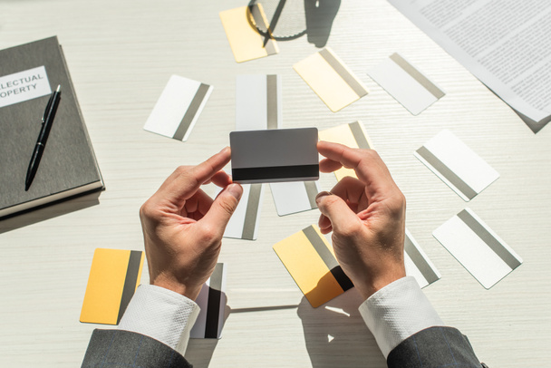 Обрезанный вид бизнесмена, держащего кредитную карту возле ноутбука и бизнес-документа на белом текстурированном фоне - Фото, изображение