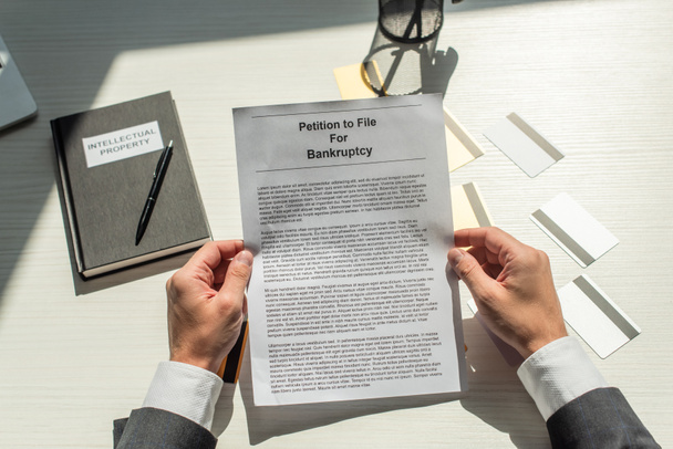 Обрезанный взгляд бизнесмена, держащего петицию о банкротстве возле кредитных карт и ноутбука на белом текстурированном фоне - Фото, изображение