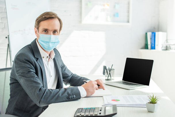 Businessman in medical mask looking at camera, while writing in notebook at workplace on blurred background - Fotoğraf, Görsel