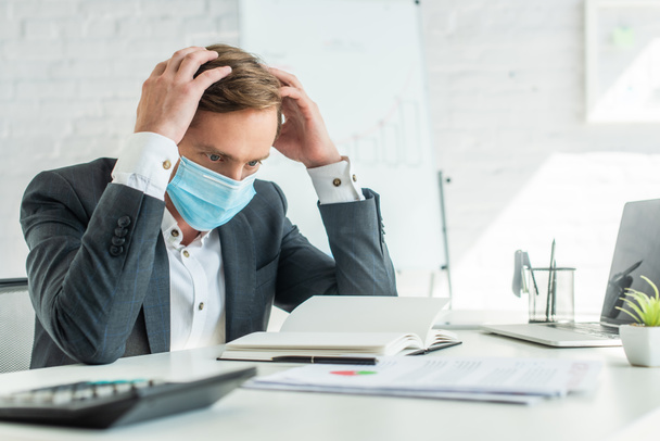 Банкрот в медицинской маске, держась за руки возле головы, сидя на рабочем месте с размытым флипчартом на заднем плане - Фото, изображение