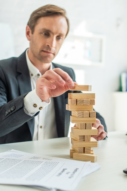 Focused businessman building blocks wood game, while sitting at workplace with blurred petition for bankruptcy on foreground - Фото, изображение