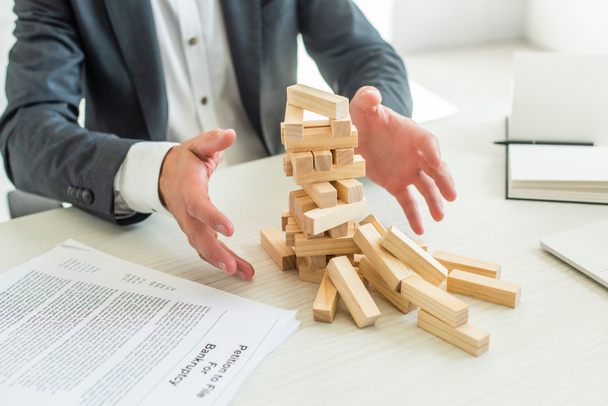 Cropped view of businessman with hands near blocks wood game falling, sitting near documents at workplace - Foto, Imagen