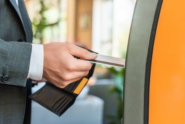 Cropped view of businessman holding wallet and credit card, while standing near atm on blurred background - Foto, immagini