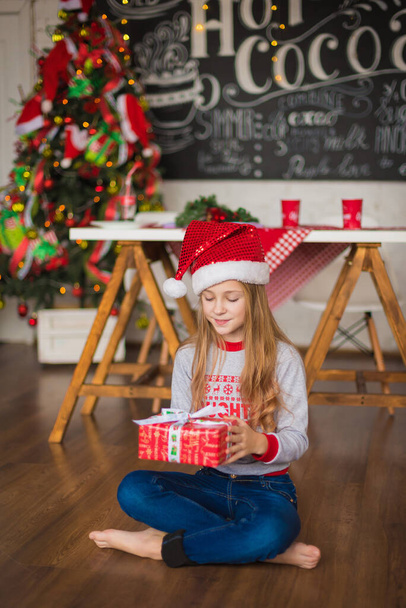 In de kerstkamer mooi jong meisje met Santa hoed op haar had het vasthouden van cadeau in haar handen. - Foto, afbeelding