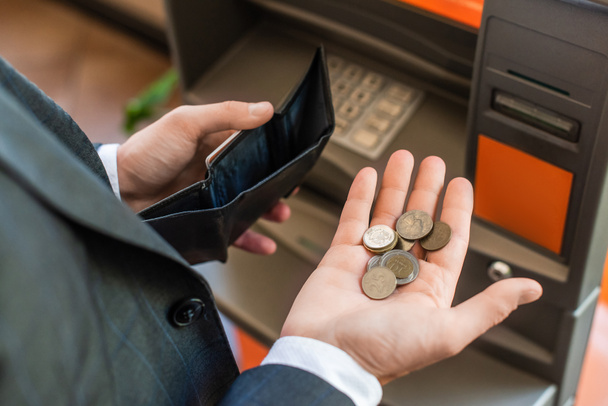 Cropped view of businessman with coins on palm, holding empty wallet, while standing near atm on blurred background - Фото, зображення
