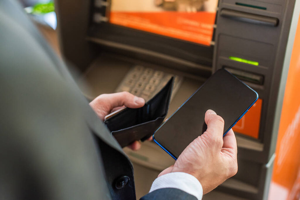 Cropped view of businessman with empty wallet, holding smartphone, while standing near atm on blurred foreground - Fotografie, Obrázek