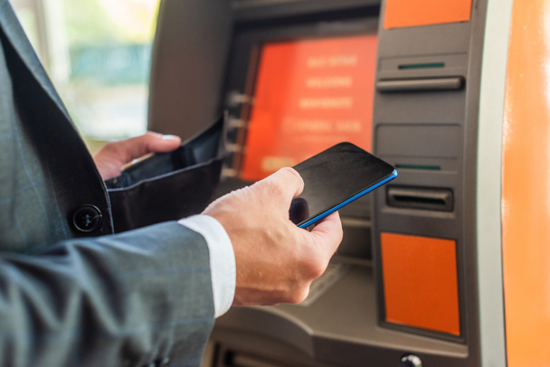 Cropped view of businessman with empty wallet and smartphone standing near atm on blurred background - Foto, immagini