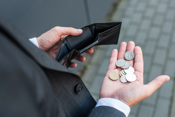 Vista cortada de pobre empresário com moedas na palma da mão, segurando carteira vazia com rua embaçada no fundo - Foto, Imagem