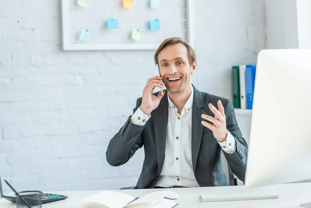Alegre gesto de hombre de negocios, mientras habla por teléfono en el lugar de trabajo con monitor de computadora borrosa en primer plano - Foto, imagen