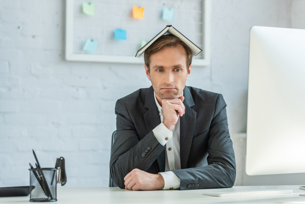 Empresario molesto con cuaderno en la cabeza, sentado en el lugar de trabajo con organizador de malla borrosa en el fondo - Foto, imagen