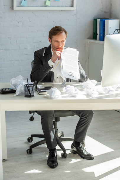 Rozrušený obchodník, který drží petici za bankrot, zatímco sedí u stolu se zmačkanými papíry v kanceláři - Fotografie, Obrázek