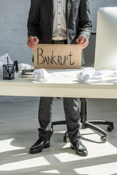 Vista recortada del empresario sosteniendo cartón con letras en bancarrota, mientras está parado en el lugar de trabajo - Foto, Imagen