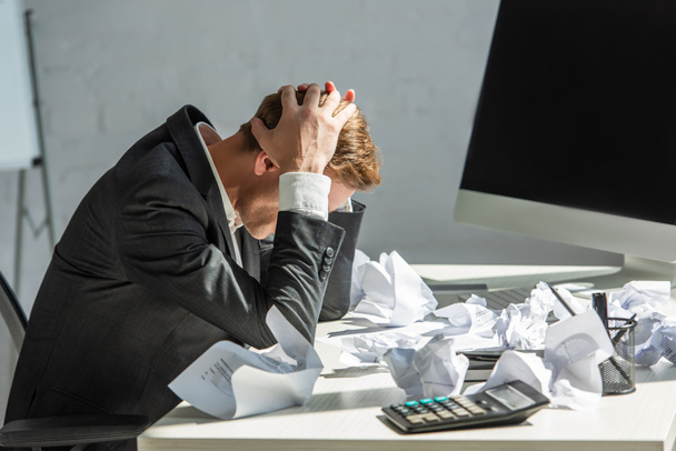 Empresario estresado con las manos en la cabeza, sentado en el lugar de trabajo con papeles desmenuzados sobre fondo borroso - Foto, Imagen