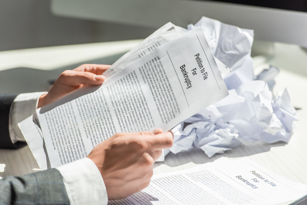 Widok przedsiębiorcy trzymającego petycję o bankructwo w pobliżu stosu pogniecionych papierów na niewyraźnym tle - Zdjęcie, obraz