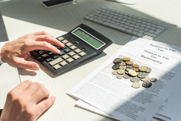 Getroffen beeld van zakenman rekenen op rekenmachine in de buurt van munten op petities voor faillissement op tafel - Foto, afbeelding