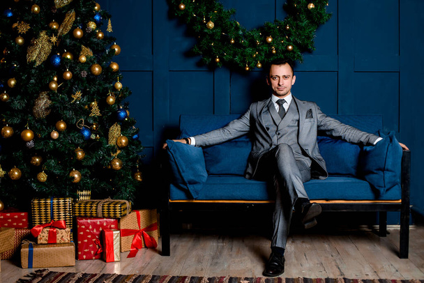 Empresário em um terno cinza no escritório na véspera de Ano Novo. Sentado em um sofá azul perto de uma árvore de Natal vermelho e caixas de presente. à espera da noite de Natal - Foto, Imagem
