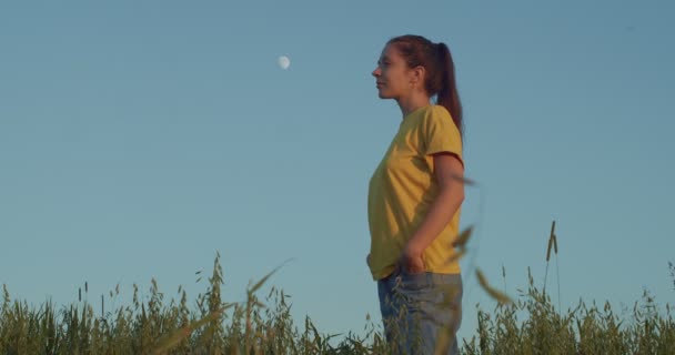 Side view of young girl standing outdoors in t-shirt thinking of something pleasant. Smiling woman dreaming of happy future on blue sky moon background slow motion. Freedom happiness enjoyment - Footage, Video