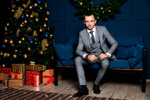 man in a gray suit in the office on New year's eve. Sitting on a blue sofa near a Christmas tree red and gift boxes. waiting for Christmas night - Foto, Imagem