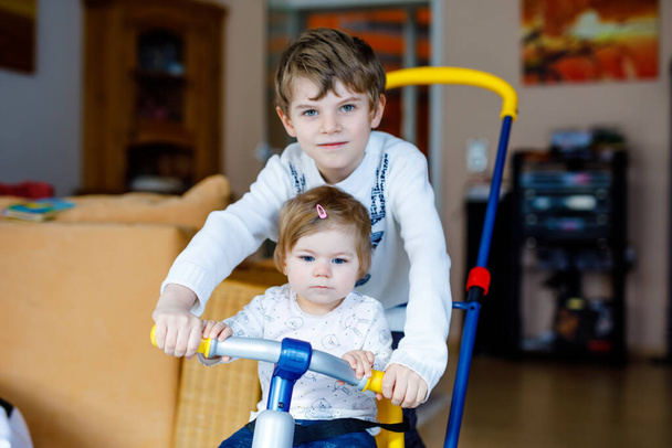 Kleine jongen die fiets of driewieler duwt met een schattig zusje. Peuter meisje en broer spelen samen thuis, binnen. Broers en zussen verbroedering en liefde, gezamenlijke en gemeenschappelijke activiteit en spel. - Foto, afbeelding