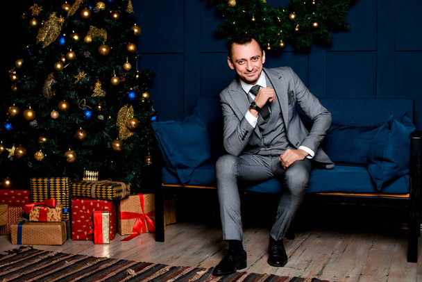 Бизнесмен в сером костюме в офисе в канун Нового года. Сидя на синем диване рядом с елкой красной и подарочными коробками. В ожидании рождественской ночи.. - Фото, изображение
