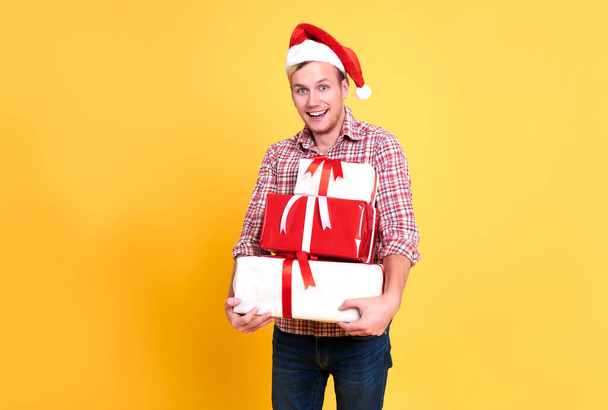 Schöner Mann trägt Weihnachtsmütze mit einer Menge Geschenkschachteln auf gelbem Hintergrund. Weihnachten und ein gutes neues Jahr Konzept. - Foto, Bild