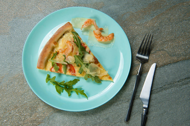 Hühnerpizza mit Zutaten auf dem Teller. Messer und Gabel in der Nähe von Fast-Food-Gericht auf anthrazitfarbenem Hintergrund. Stück Meeresfrüchte-Pizza mit Garnelen, frischen Rucolablättern und geschnittenem Parmesan - Foto, Bild