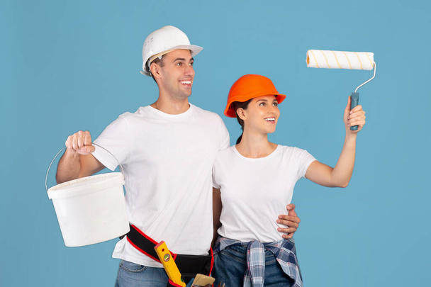 Happy young couple in hard hats with paint bucket and roller brush - Photo, Image
