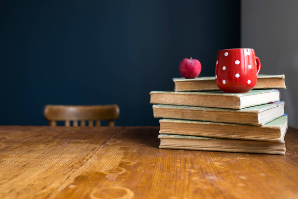 A stack of large, old, rag-bound books lie on a wooden table against a dark blue wall. Pen and notebook. Concept - back to school. Online training. Copy space. Select focus. Red cup with white polka dots with tea. - 写真・画像