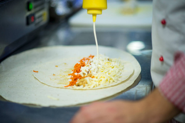 Kokki tekee kananlihan shawarmaa ammattimaisessa ravintolakeittiössä. Lavash-leipä (pita) porkkanalla ja juustolla. Gyros ruoanlaitto prosessi. Kokki kaatoi majoneesia pullosta. Liha Tortilla - Valokuva, kuva