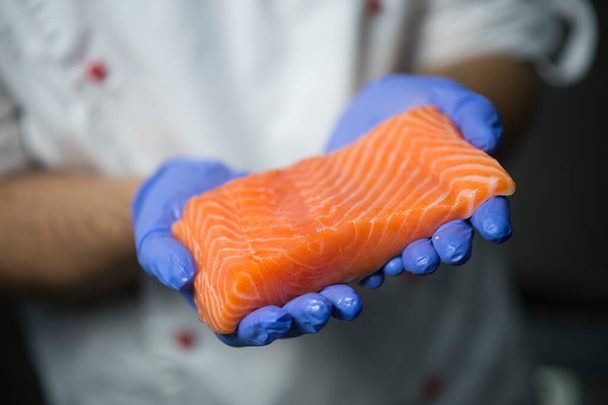 Sushi Chef tenant un filet de saumon cru frais dans les mains dans des gants de protection. Filet de poisson en bref sur fond flou. Sushi maître tient le poisson avant la cuisson des sushis - Photo, image